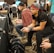 Several people are sitting in a room with gaming consoles and monitors, focusing intently on their screens. The setting appears to be a casual gaming event, with participants holding controllers while playing video games. The room has a casual atmosphere with chairs and tables set up for gaming.