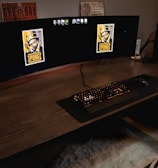 A gaming setup featuring a large curved monitor displaying PUBG logos on the screens. A keyboard and mouse are positioned on a large desk pad. A pair of headphones rests on the desk alongside a mobile phone. A drink in a Starbucks cup is placed to the right, while a desktop tower is seen in the background on the right side.