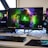 A gaming setup featuring a central monitor with a vibrant display showing 'The Spirit of Gaming'. On either side, there are gaming PCs with illuminated components and transparent cases. Headsets hang from the side of the monitor. The desk also has a black keyboard and mouse, and a branded mousepad.