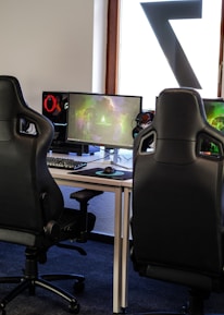 A row of gaming stations equipped with large monitors, gaming chairs, keyboards, and headphones. The setup is organized and ready for gaming, featuring a colorful screen display. The room has a modern and tech-savvy ambiance.