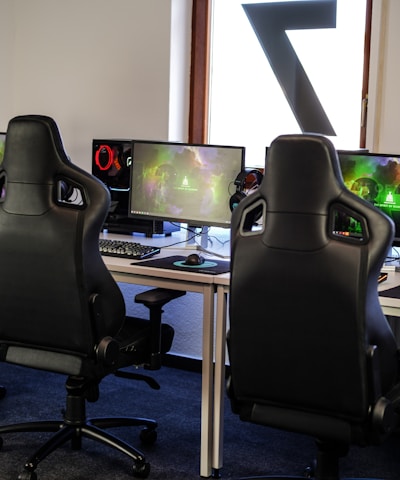 A row of gaming stations equipped with large monitors, gaming chairs, keyboards, and headphones. The setup is organized and ready for gaming, featuring a colorful screen display. The room has a modern and tech-savvy ambiance.