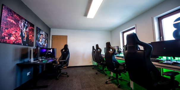 A well-lit gaming room features multiple gaming stations with high-backed chairs and monitors. The room has a minimalist setup with green and black color tones. Large posters adorn the walls, and the room appears clean and organized.