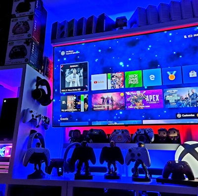A modern gaming setup featuring a large wall-mounted screen displaying a gaming console interface. The room is decorated with gaming-themed items such as controllers and collectibles. The ambient lighting includes shades of blue and red, creating a vibrant atmosphere.