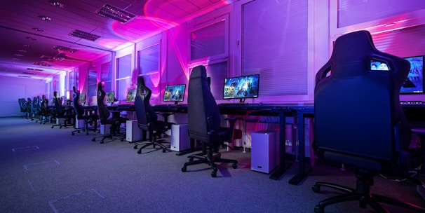 A row of gaming computers with large monitors and racing-style chairs is set up along the interior of a modern room. The space is illuminated with vibrant purple and blue LED lighting, creating a futuristic atmosphere. The room is organized and clean, suggesting a professional gaming or esports environment.