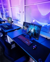 A row of modern gaming setups is arranged neatly along a window-lined wall. Each station includes a high-resolution monitor displaying vivid graphics, a mechanical keyboard with colored lighting, a gaming mouse, and an ergonomic chair. The room is illuminated with ambient blue and purple lights, creating a sleek and futuristic atmosphere.