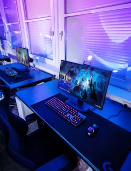 A row of modern gaming setups is arranged neatly along a window-lined wall. Each station includes a high-resolution monitor displaying vivid graphics, a mechanical keyboard with colored lighting, a gaming mouse, and an ergonomic chair. The room is illuminated with ambient blue and purple lights, creating a sleek and futuristic atmosphere.