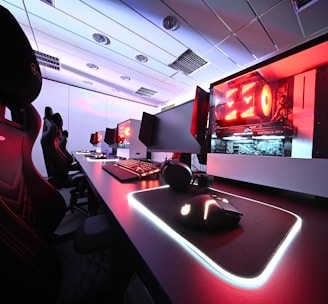 A row of high-end gaming computers on sleek desks, featuring RGB lighting and gaming accessories. The computers are housed in transparent cases with visible components, emitting a red glow. Comfortable gaming chairs with ergonomic design are positioned next to the desks, and the room's ceiling is modern with recessed lighting.