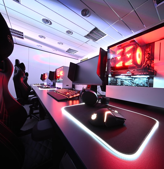A row of high-end gaming computers on sleek desks, featuring RGB lighting and gaming accessories. The computers are housed in transparent cases with visible components, emitting a red glow. Comfortable gaming chairs with ergonomic design are positioned next to the desks, and the room's ceiling is modern with recessed lighting.
