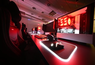 A modern gaming setup featuring several high-end computers with illuminated red interior lighting. The room is equipped with gaming chairs exhibiting a sleek black and red design. Multiple monitors are arranged on a long desk, and peripheral devices like keyboards and mice are uniformly placed, reflecting an ambiance ideal for gaming or competitive play.
