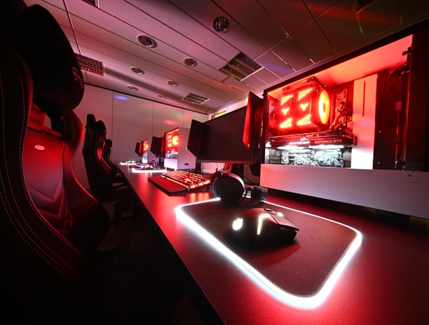 A modern gaming setup featuring several high-end computers with illuminated red interior lighting. The room is equipped with gaming chairs exhibiting a sleek black and red design. Multiple monitors are arranged on a long desk, and peripheral devices like keyboards and mice are uniformly placed, reflecting an ambiance ideal for gaming or competitive play.