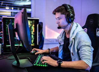 A young person is intensely focused on a computer screen while wearing a headset with a microphone. They are using a gaming keyboard and mouse, sitting in a gaming chair. The room has cool lighting, with a visible PC tower.