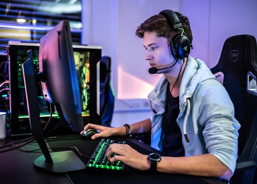 A young person is intensely focused on a computer screen while wearing a headset with a microphone. They are using a gaming keyboard and mouse, sitting in a gaming chair. The room has cool lighting, with a visible PC tower.