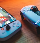 A gaming scenario featuring a smartphone with a game interface next to two controllers on a wooden surface. The smartphone displays vibrant images and icons, while the controllers feature a sleek, modern design with buttons and joysticks prominently visible. Warm lighting enhances the scene.