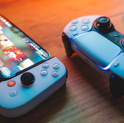 A gaming scenario featuring a smartphone with a game interface next to two controllers on a wooden surface. The smartphone displays vibrant images and icons, while the controllers feature a sleek, modern design with buttons and joysticks prominently visible. Warm lighting enhances the scene.