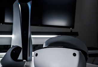A sleek virtual reality headset sits on a reflective surface beside a gaming console and its controller. The background features a large dark monitor screen, creating a modern and futuristic setting. The lighting is subdued, highlighting the glossy surfaces of the electronic devices.