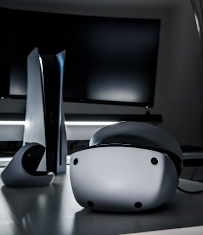 A sleek virtual reality headset sits on a reflective surface beside a gaming console and its controller. The background features a large dark monitor screen, creating a modern and futuristic setting. The lighting is subdued, highlighting the glossy surfaces of the electronic devices.