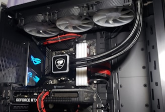 A high-performance gaming computer setup showing the interior components, including a GeForce RTX graphics card, cooling system with three fans, a motherboard, and visible wiring with red and black cables.