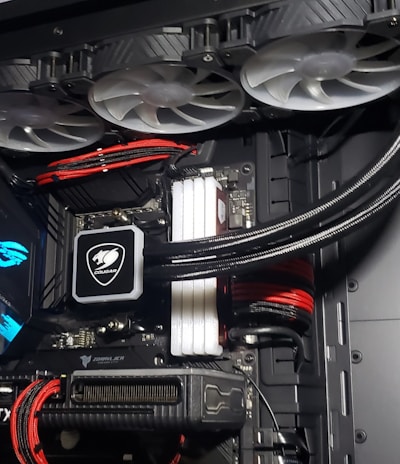 A high-performance gaming computer setup showing the interior components, including a GeForce RTX graphics card, cooling system with three fans, a motherboard, and visible wiring with red and black cables.