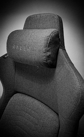 A high-quality, fabric gaming chair featuring ergonomic design and enhanced comfort. The chair has a distinct headrest with visible stitching, embossed branding, and a modern aesthetic.