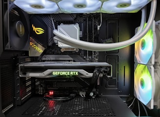 Inside view of a computer case showcasing a sophisticated gaming setup. Visible components include a GeForce RTX graphics card illuminated with green lighting, an ASUS motherboard, cooling fans with LED lighting, and a liquid cooling system with white tubes. Cables are neatly organized, and overall, the hardware suggests a high-performance machine.
