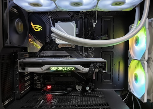 Inside view of a computer case showcasing a sophisticated gaming setup. Visible components include a GeForce RTX graphics card illuminated with green lighting, an ASUS motherboard, cooling fans with LED lighting, and a liquid cooling system with white tubes. Cables are neatly organized, and overall, the hardware suggests a high-performance machine.
