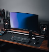 A modern gaming and streaming setup with a curved monitor placed centrally on a wooden desk. Surrounding the monitor are powerful speakers and a high-end PC tower with glowing fans. Various peripherals, including a mechanical keyboard, ergonomic mouse, and audio mixer, are neatly arranged. Overhead, a professional microphone and stand are ready for use, possibly for gaming commentary or podcasting.