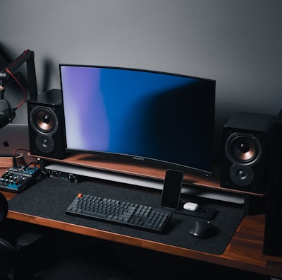A modern gaming and streaming setup with a curved monitor placed centrally on a wooden desk. Surrounding the monitor are powerful speakers and a high-end PC tower with glowing fans. Various peripherals, including a mechanical keyboard, ergonomic mouse, and audio mixer, are neatly arranged. Overhead, a professional microphone and stand are ready for use, possibly for gaming commentary or podcasting.