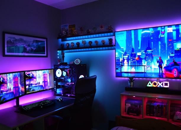 A vibrant gaming setup featuring multiple monitors displaying a cyberpunk cityscape. The room is illuminated with neon purple and blue LED lights, adding a futuristic ambiance. A custom-built PC with glowing components sits to the side, and shelves with collectible figurines are visible. A large digital screen on the wall shows an animated city scene, enhancing the immersive atmosphere.